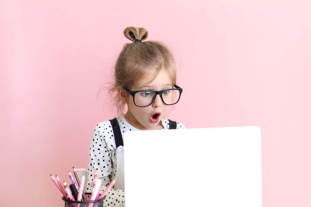Auf dem Bild ist ein kleines Mädchen mit großer dunkler Brille zu sehen, dass an einem Laptop sitzt und ein überraschtes Gesicht macht. Der Hintergrund im Bild ist pink.