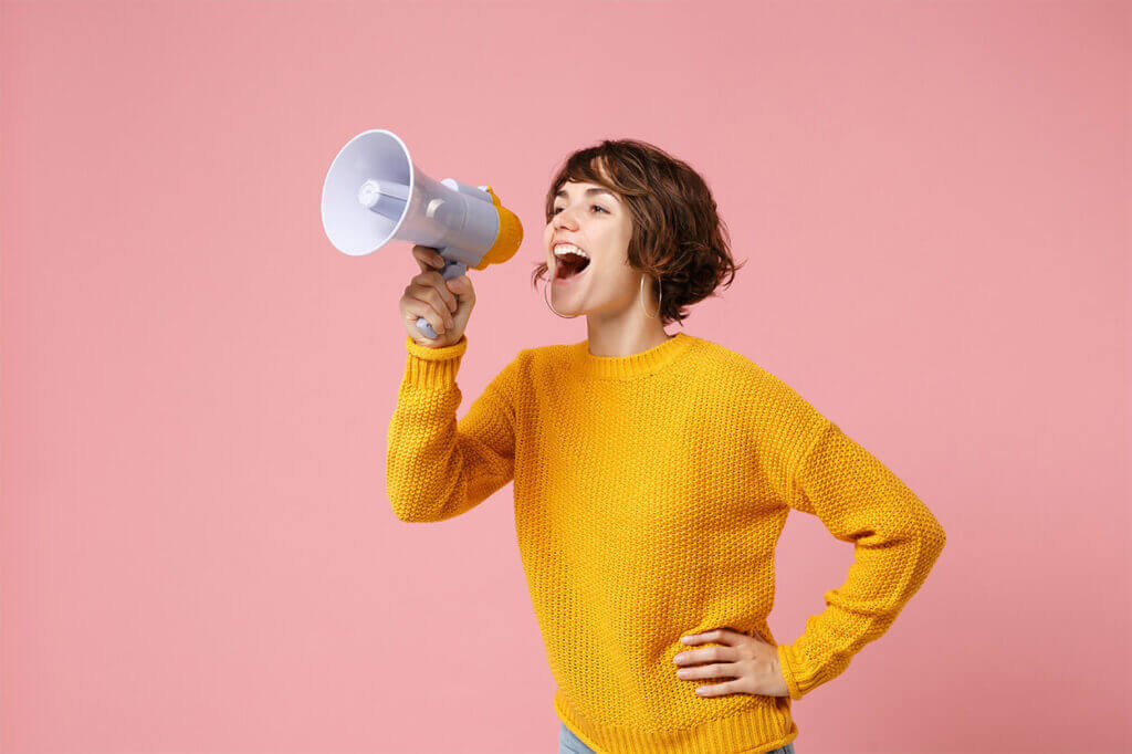In order for subscription business to be successful, you need the right salespeople. The picture shows a young woman with short brown hair and a yellow sweater. The woman is holding a megaphone through which she is shouting. The background of the picture is colored corall.