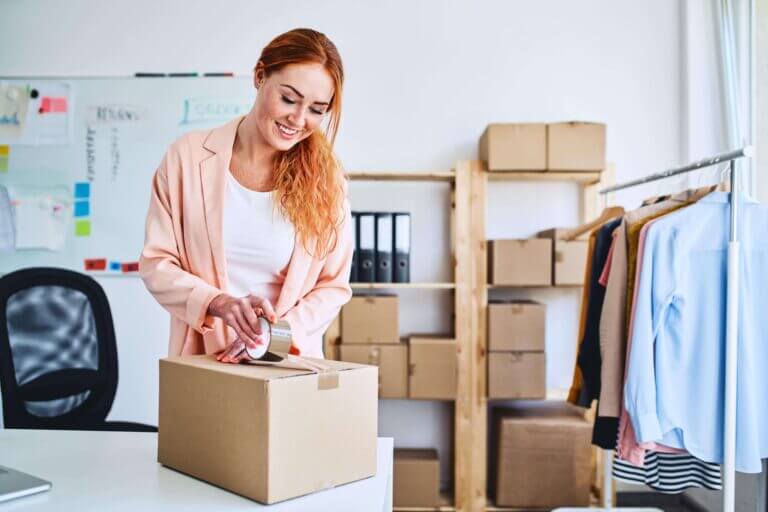 Eine Frau mit roten Haaren und rosafarbenem Blaser steht in ihrem Büro und verpackt Waren, die sie mit der Post verschicken will.