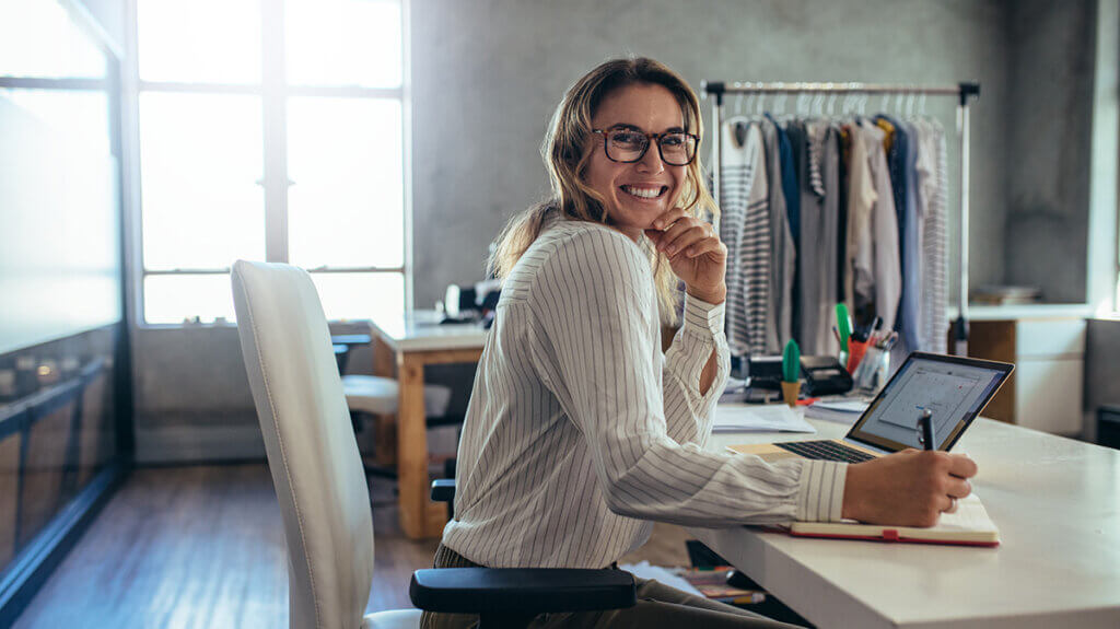 Abos im Trend. Das Bild zeigt eine Frau in ihrem Büro. Sie sitzt an ihrem Schreibtisch und arbeitet am PC. Sie blick lächelnd zur Kamera. Im Hintergrund sind Verkaufsartikel, wie Kleidung zu sehen.