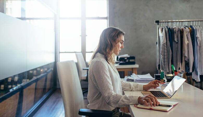 The picture shows a businesswoman in her office working on her laptop. Various items of clothing are hung in the background.